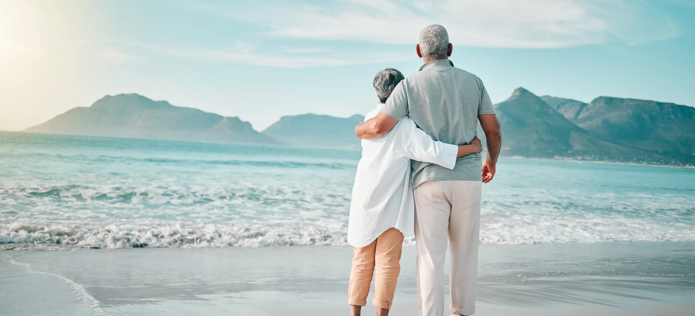 Retired couple standing arm in arm on a quiet beach, looking out at the ocean and distant mountains.